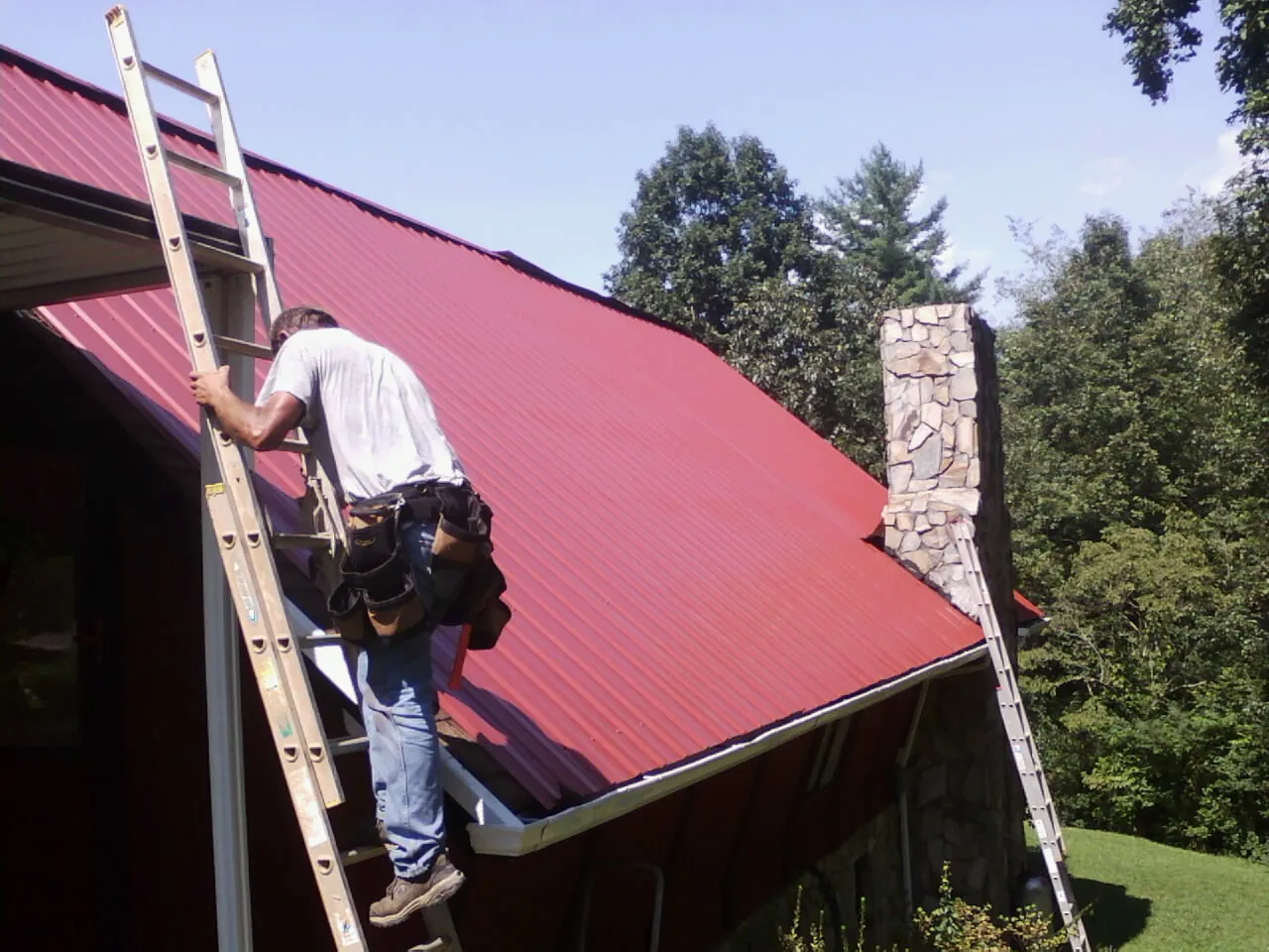 Roofer on a ladder beside a red metal roof and stone chimney — Soffit & Fascia Repair by Roofing Brothers in Wade
